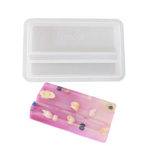 May include: A clear rectangular silicone mold with two compartments and the text "V090-3". Below is a pink resin phone stand decorated with seashells, pearls, and blue and black beads. The stand has two slots.