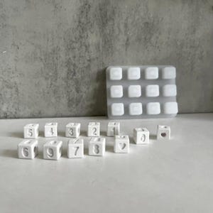 Op de afbeelding: Witte keramische nummer kralen met een hartkraal. De kralen zijn in een rij op een wit oppervlak gerangschikt. Er is een heldere siliconen mal op de achtergrond.
