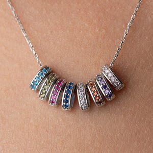 May include: A silver necklace featuring a delicate chain and a series of colorful, gem-studded rings. The rings display a spectrum of colors, including blue, pink, and white, creating a vibrant and eye-catching design. The necklace is worn around the neck.