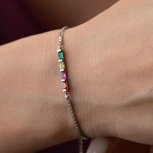 Può includere: Un braccialetto d'argento con una catena delicata e una fila di pietre preziose rettangolari colorate. Le pietre preziose includono verde, giallo, rosa e rosso. Il braccialetto è su un polso.