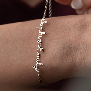 May include: A silver bracelet featuring the names "Sophia" and "Emily" in a cursive font. The bracelet is suspended from a fine chain and is displayed against a wrist. The background is a blurred maroon hue.