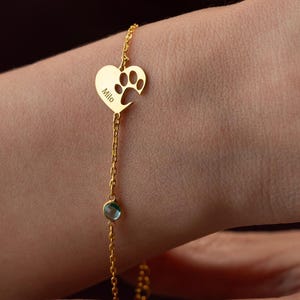 May include: A gold-colored bracelet featuring a heart-shaped charm with a paw print and the name "Milo" engraved. The bracelet has a delicate chain and a small, round, blue gemstone. The bracelet is worn on a wrist.
