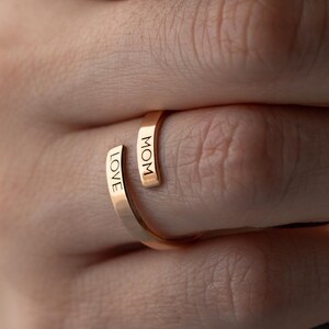 May include: A gold-colored ring with the words "LOVE" and "MOM" engraved on it. The ring is wrapped around a finger, showcasing the delicate design and the message of affection. The ring is a piece of jewelry.