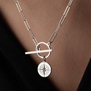 May include: Silver-toned necklace with a compass pendant. The necklace features a toggle clasp and a chain with rectangular links. The compass pendant is round with a detailed design. The necklace is displayed against a dark background.