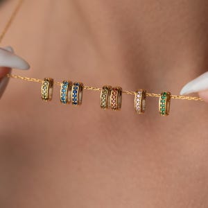 May include: A gold necklace featuring multiple small, colorful, gemstone-studded rings. The rings are in shades of green, blue, and gold. The necklace is held by a person with white nail polish.