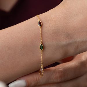 Könnte beinhalten: Ein zartes goldenes Armband mit drei ovalen Edelsteinen in Rot, Grün und Klar. Das Armband wird am Handgelenk getragen und zeigt die feine Kette und die farbenfrohen Edelsteine.