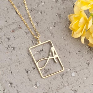 Peut inclure: Un collier doré avec un pendentif rectangulaire orné de la lettre "A" en cursive. Le collier est présenté sur une surface grise texturée, avec un bouquet de fleurs jaunes en arrière-plan.