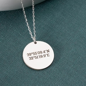 Puede incluir: Un collar plateado con un colgante redondo. El colgante tiene coordenadas grabadas: 39°55'09.4"N 32°51'19.9"E. El collar se muestra sobre una superficie blanca sobre un fondo verde azulado.