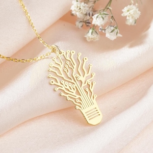 Pode incluir: Um colar de ouro com um pingente de lâmpada que apresenta um design de placa de circuito. O colar está sobre um fundo de tecido rosa.