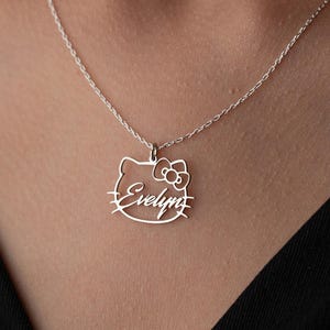 May include: A silver necklace with a pendant in the shape of a cartoon cat's head, complete with a bow. The name "Evelyn" is written in cursive script below the cat's face. The necklace is set against a dark backdrop.