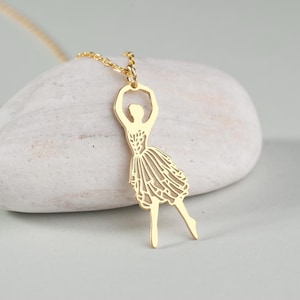Può includere: Una collana d'oro con un delicato ciondolo a forma di ballerina. La ballerina è in una posa di danza con le braccia alzate sopra la testa.