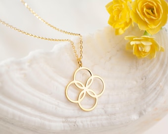 May include: A delicate gold necklace with a four-circle pendant. The pendant is centered on a fine gold chain. The necklace rests on a white, textured surface, with yellow flowers in the background.