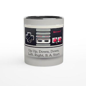 May include: A light gray ceramic mug featuring a black and gray video game controller design. The controller has red buttons and the text "Up Up, Down, Down, Left, Right, B, A, Start" is printed below the controller.