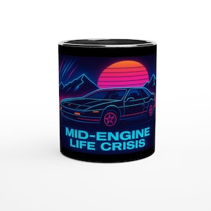 Op de afbeelding: Zwarte koffiemok met een retro design met een autosilhouet, bergen en een zonsondergang. De mok heeft de tekst "MID-ENGINE LIFE CRISIS" in blauwe neonletters. De mok heeft een zwart interieur en rand.
