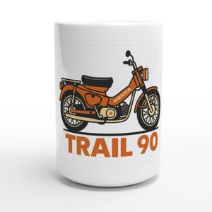 May include: White ceramic mug with an orange illustration of a Honda Trail 90 motorcycle. The mug has the words "TRAIL 90" in orange block letters below the motorcycle graphic. The motorcycle is detailed with black and cream accents.