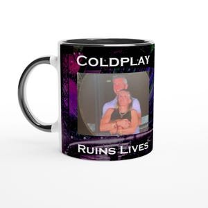 May include: White ceramic mug with a black interior and handle. The mug features the text "COLDPLAY" and "RUINS LIVES" in white, along with a photo of two people. The background has a purple and green design.