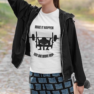 May include: A woman wearing a white t-shirt with the text "MAKE IT HAPPEN" and a black silhouette of a person weightlifting with the text "JUST ONE MORE REP" underneath. She is also wearing black athletic shorts with a repeating pattern of the text "UPLOAD YOUR IMAGE" and a black jacket.