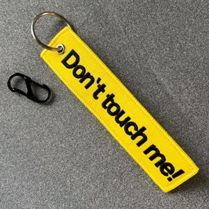 以下が含まれることがあります： 黄色い布製のキーホルダーに黒い刺繍で「Don't touch me!」と書かれている