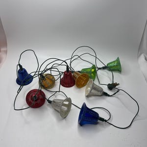 May include: A string of colorful bell-shaped Christmas lights. The lights are in various colors, including red, blue, green, yellow, and silver. The lights are connected by a black wire and have a standard plug.