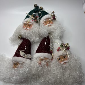 May include: A collection of Santa Claus head ornaments. Each ornament features a detailed face with glasses, a fluffy white beard, and a hat in either green or burgundy. The hats are adorned with small bells and decorative accents.