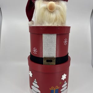 May include: A red Christmas-themed gift box set with a Santa Claus figure on top. The boxes are decorated with snowflakes, Christmas trees, and a gift box. The Santa figure has a white beard and a burgundy hat.