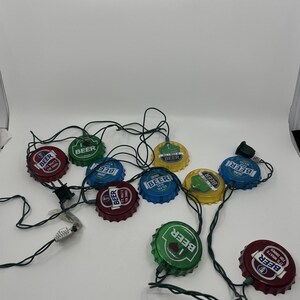 May include: A string of decorative beer bottle cap lights in various colors, including red, green, blue, and yellow. Each cap has the word "BEER" printed on it, with additional text. The lights are connected by a black wire.