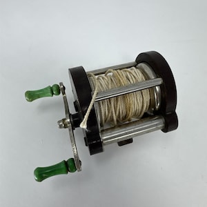 May include: Vintage fishing reel with a dark brown frame, silver metal components, and green handles. The reel is filled with off-white fishing line. A classic piece of fishing equipment.