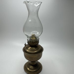May include: An antique oil lamp with a clear glass chimney and a brass base. The chimney has a ruffled top. The lamp's base is round and has a classic design. The lamp is designed to hold oil for illumination.