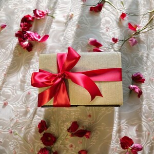 May include: A gift box wrapped in beige paper with a large red satin bow. The box is surrounded by pink flower petals.