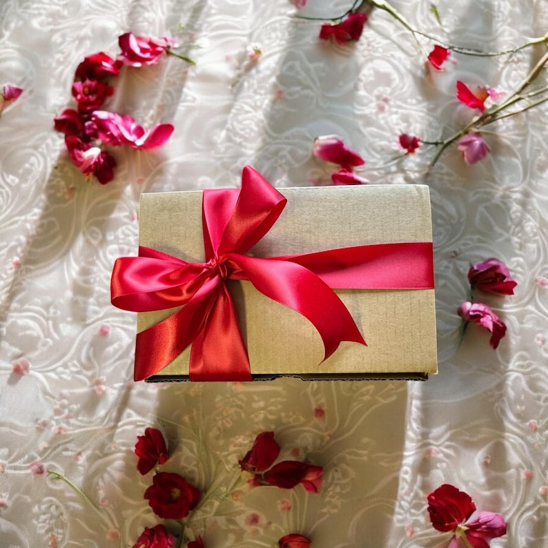 May include: A gift box wrapped in beige paper with a large red satin bow. The box is surrounded by pink flower petals.