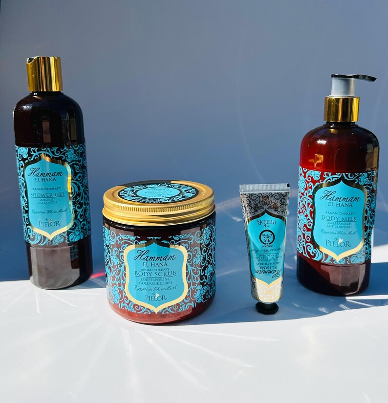 May include: A set of four bath and body products in a Moroccan-inspired design. The products include a shower gel, body scrub, body milk, and hand cream. The bottles and jars are all brown with gold accents and feature a blue and white pattern. The products are labeled "Hammam El Hana" and "PIELOR".