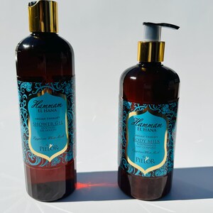 May include: Two amber glass bottles with gold lids and blue and gold labels. The bottles contain shower gel and body milk. The labels feature the text "Hammam El Hana" and "Argan Therapy".