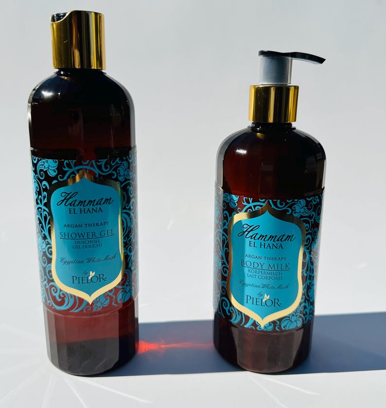 May include: Two amber glass bottles with gold lids and blue and gold labels. The bottles contain shower gel and body milk. The labels feature the text "Hammam El Hana" and "Argan Therapy".