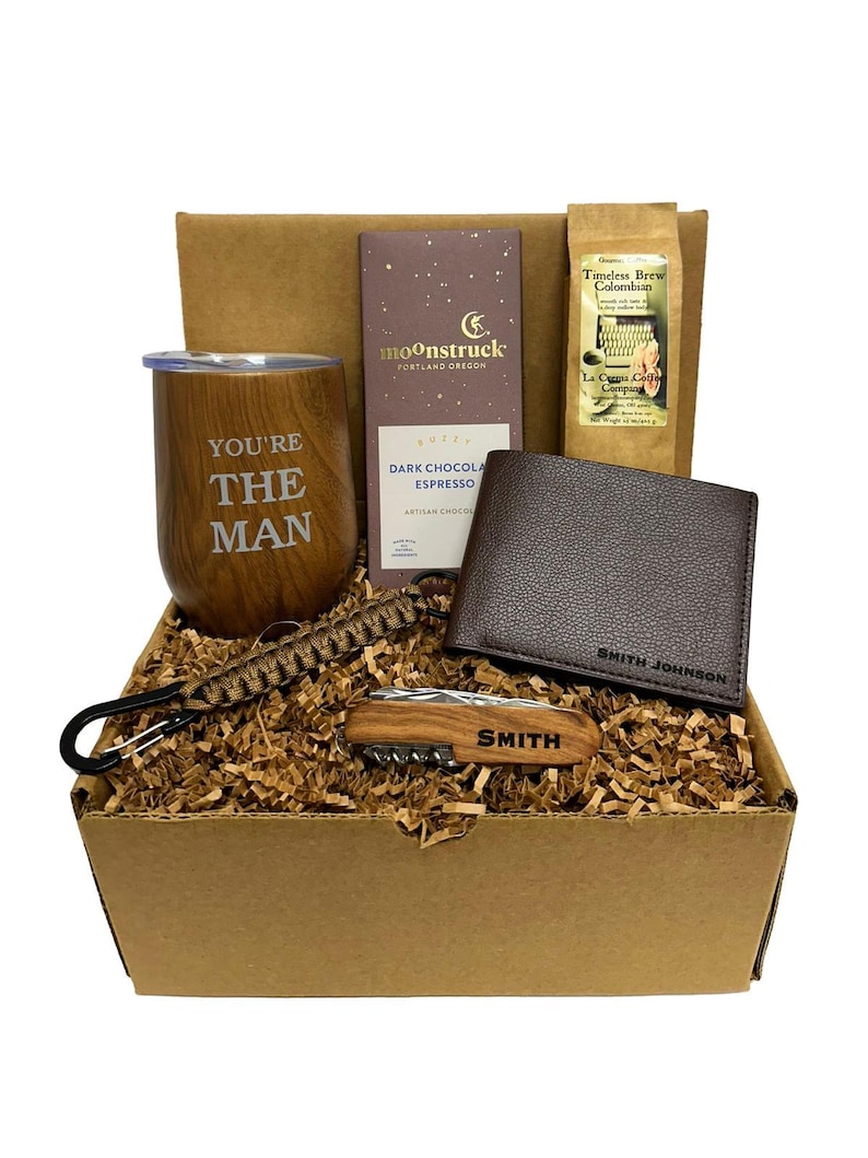 May include: A brown cardboard gift box filled with a variety of items, including a brown leather wallet, a stainless steel tumbler with the words "You're The Man" engraved on it, a small wooden multi-tool, a bar of dark chocolate espresso, and a bag of coffee beans.