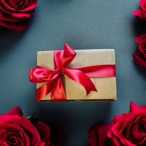 May include: A brown gift box with a red satin ribbon tied in a bow. The box is surrounded by red roses.