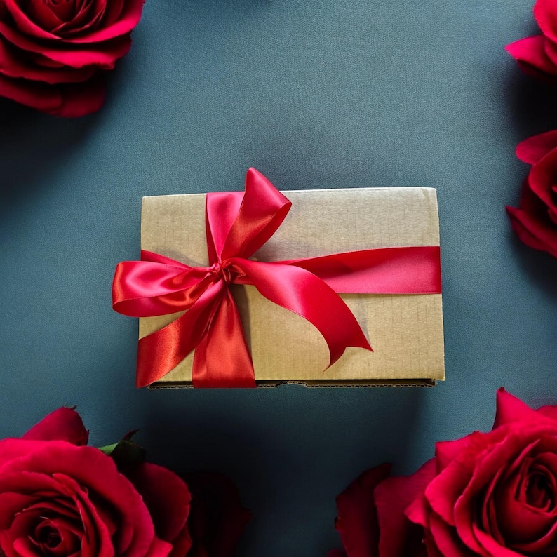 May include: A brown gift box with a red satin ribbon tied in a bow. The box is surrounded by red roses.