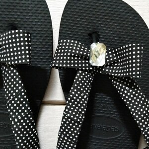 May include: Close-up of a pair of black flip-flops with white polka dot straps. The straps are adorned with a bow and a clear, faceted gem. The flip-flops have a textured surface.