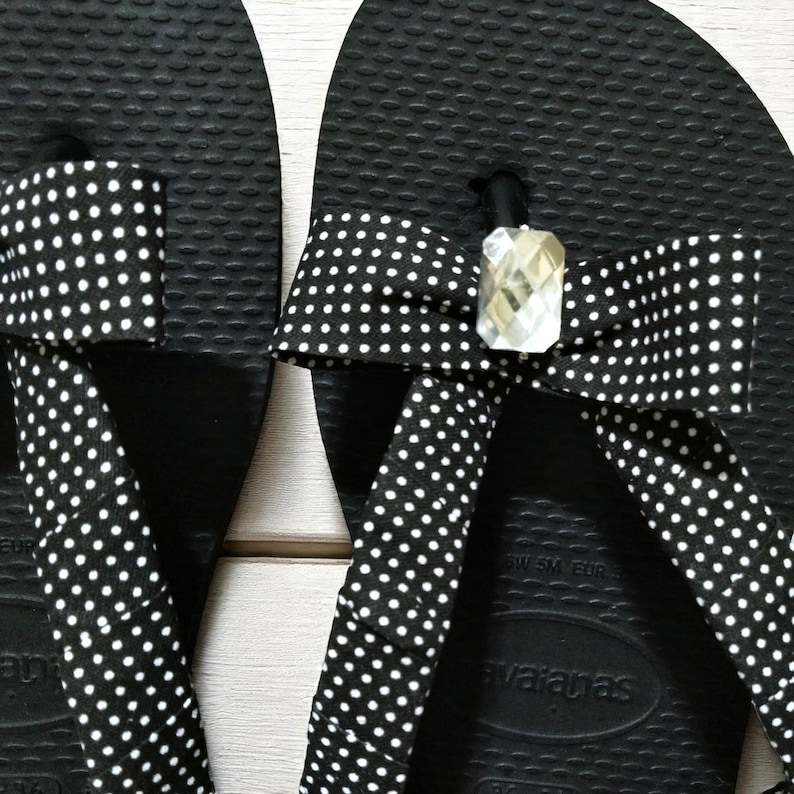 May include: Close-up of a pair of black flip-flops with white polka dot straps. The straps are adorned with a bow and a clear, faceted gem. The flip-flops have a textured surface.