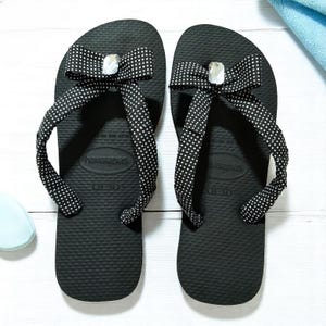 May include: A pair of black Havaianas flip-flops with white polka dot bows and a decorative gem. The flip-flops are size 35-36.