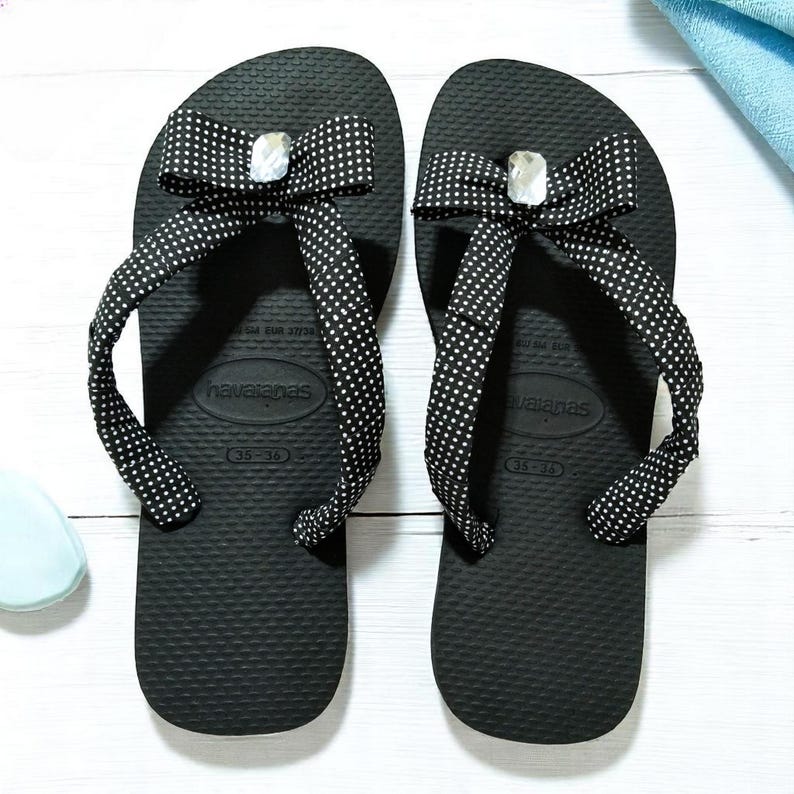 May include: A pair of black Havaianas flip-flops with white polka dot bows and a decorative gem. The flip-flops are size 35-36.