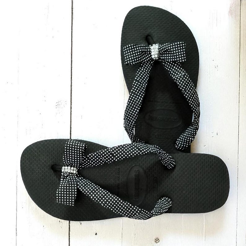 Pu&ograve; includere: Un paio di infradito nere con cinturini e fiocchi a pois bianchi. Ogni fiocco presenta un piccolo abbellimento scintillante. I sandali sono esposti su una superficie di legno bianco, mostrando un design casual ma elegante.