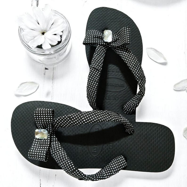 May include: Black Havaianas flip-flops with white polka dot bows and a sparkling gem accent. The sandals are displayed on a white surface with a small glass jar of white flowers.