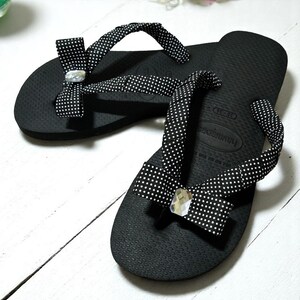 May include: Black flip-flops with white polka dot straps and decorative bows. Each bow features a clear, faceted gem. The flip-flops are on a white wooden surface, showcasing a casual footwear design.