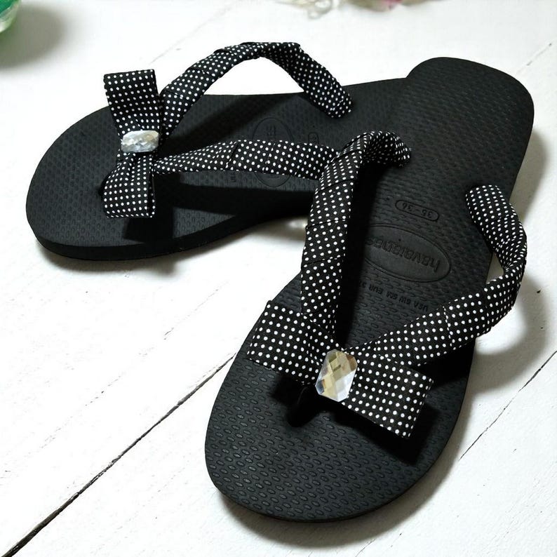 May include: Black flip-flops with white polka dot straps and decorative bows. Each bow features a clear, faceted gem. The flip-flops are on a white wooden surface, showcasing a casual footwear design.