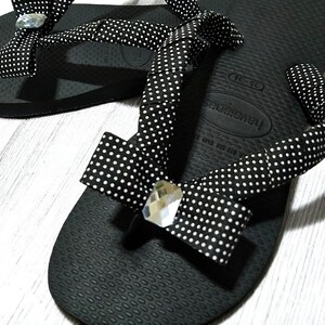May include: Black Havaianas flip-flops with white polka dot fabric straps and decorative bows. Each bow features a clear, faceted gem. The flip-flops are designed for comfort and style.