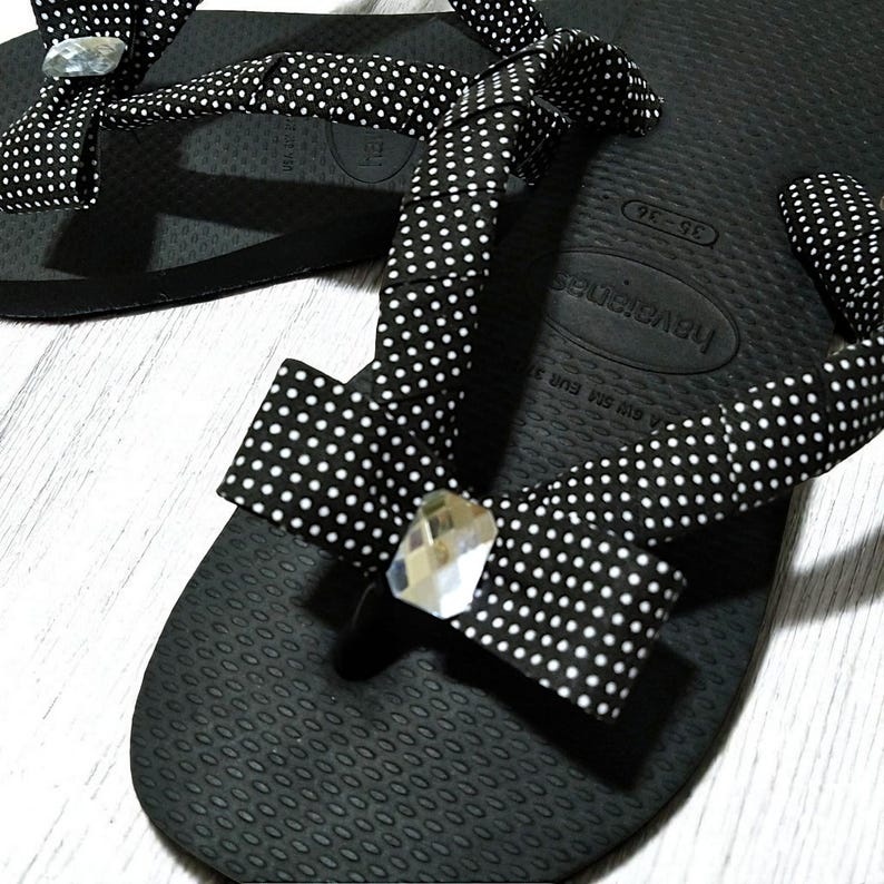 May include: Black Havaianas flip-flops with white polka dot fabric straps and decorative bows. Each bow features a clear, faceted gem. The flip-flops are designed for comfort and style.