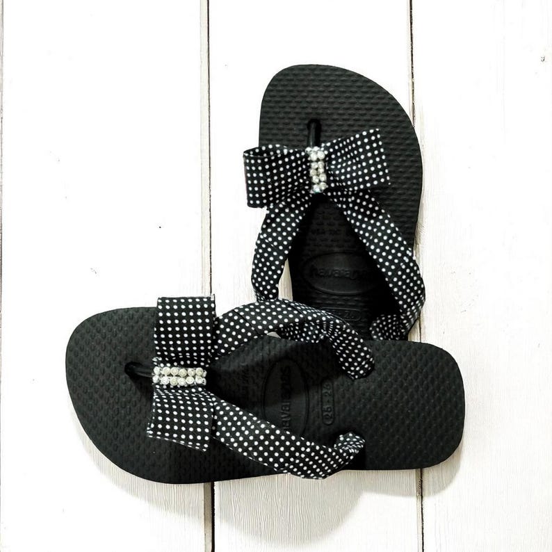Pu&ograve; includere: Infradito neri con fiocchi a pois bianchi e applicazioni di strass. I sandali sono su una superficie di legno bianco. Questi infradito sono un articolo di calzature estive.