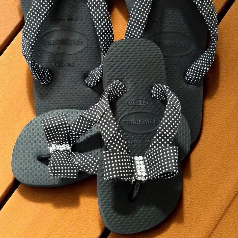 Pu&ograve; includere: Infradito neri con pois bianchi sui cinturini. Un paio presenta un fiocco con un dettaglio in strass. Gli infradito sono disposti su una superficie di legno.