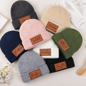 Puede incluir: Varios gorros personalizados en colores como azul marino, rosa y gris. Cada gorro tiene un parche rectangular de cuero con un nombre grabado. Los gorros están colocados sobre una superficie clara.