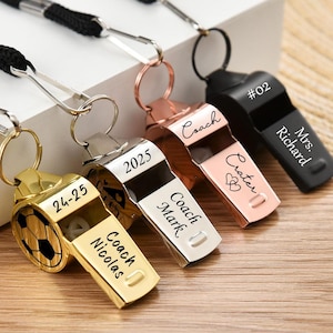 Op de afbeelding: Vijf verschillende fluitjes met sleutelhangers met gepersonaliseerde gravures. De fluitjes zijn goud, zilver, rosé goud en zwart. De gravures omvatten "Coach Crater", "Coach Mark", "Coach Nicolas", "24-25" en "Mrs. Richard #02".